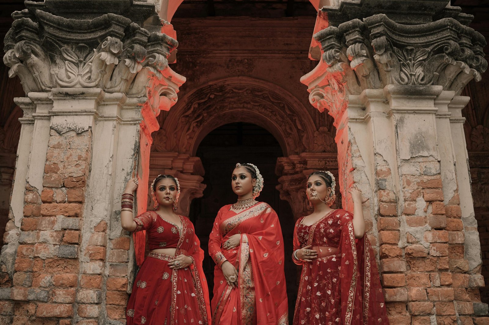 Three women in vibrant red sarees pose elegantly by ancient architecture.